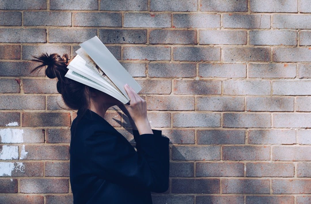 A person standing against a brick wall holding an open book across their face