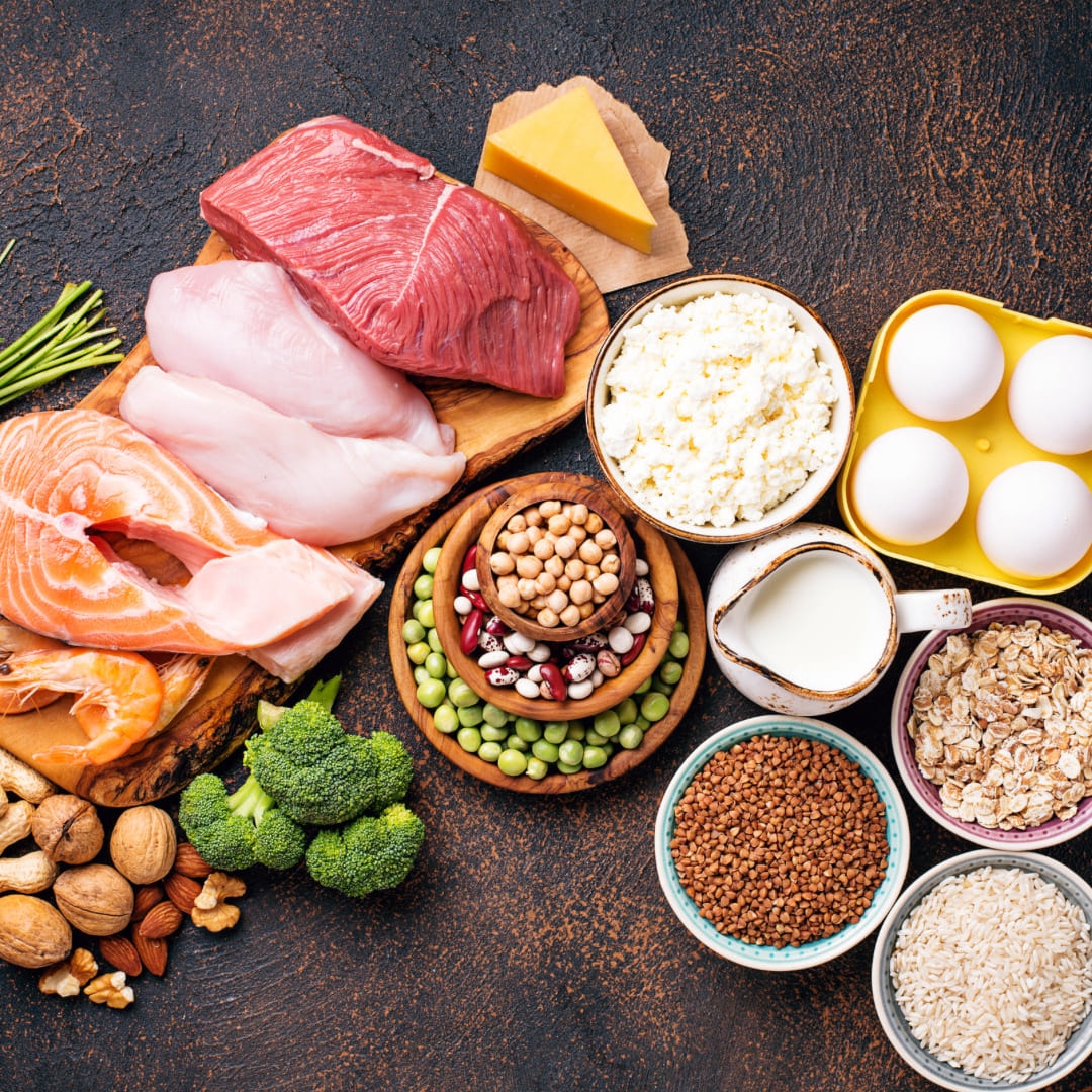 Raw steak, chicken, salmon, eggs, pulses and vegetables laid out on a dark surface